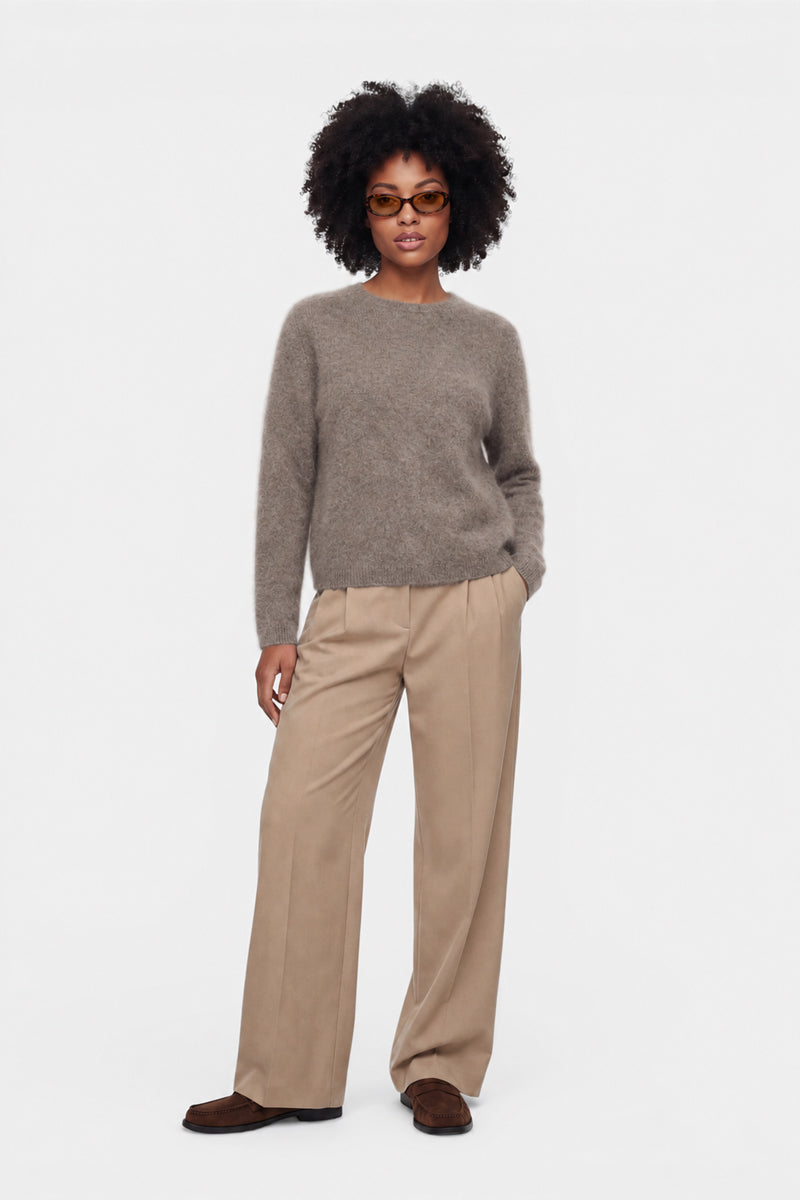 Eine Person mit kurzen Locken trägt eine braune Sonnenbrille, einen flauschigen Pullover mit Rundhalsausschnitt, eine beige Hose mit weitem Bein und braune Schuhe. Der flauschige, taupefarbene Pullover aus gebürsteter Wolle verleiht ihrem lässigen Look zusätzliche Struktur, während sie vor einem schlichten weißen Hintergrund steht, eine Hand in der Tasche hat und eine entspannte Haltung einnimmt.