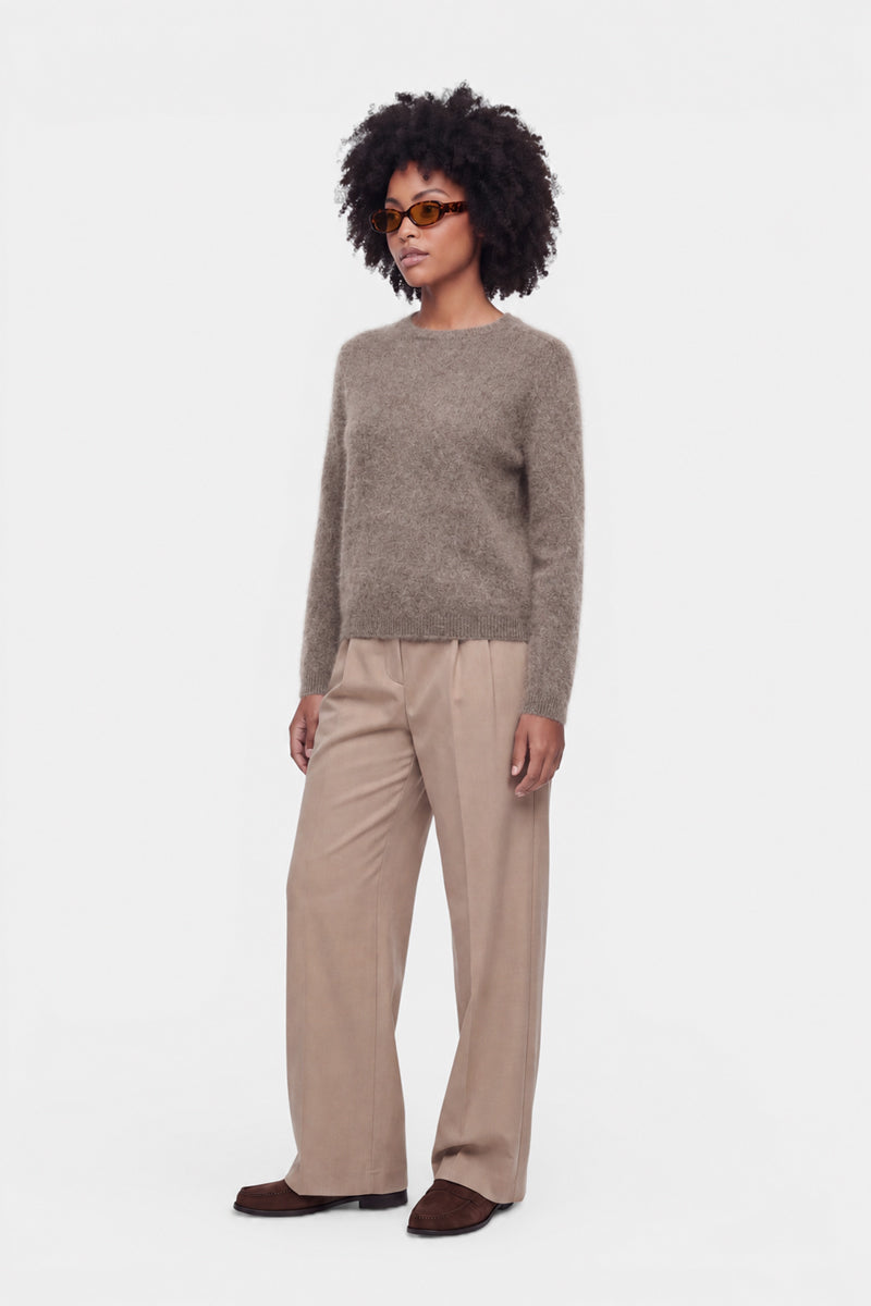 Eine Frau mit lockigem Haar trägt eine braune Sonnenbrille, einen flauschigen Pullover mit Rundhalsausschnitt aus gebürsteter Wolle, eine beige Hose mit weitem Bein und braune Schuhe. Sie steht vor einem schlichten weißen Hintergrund, blickt leicht zur Seite und lässt die Arme entspannt an den Seiten hängen.