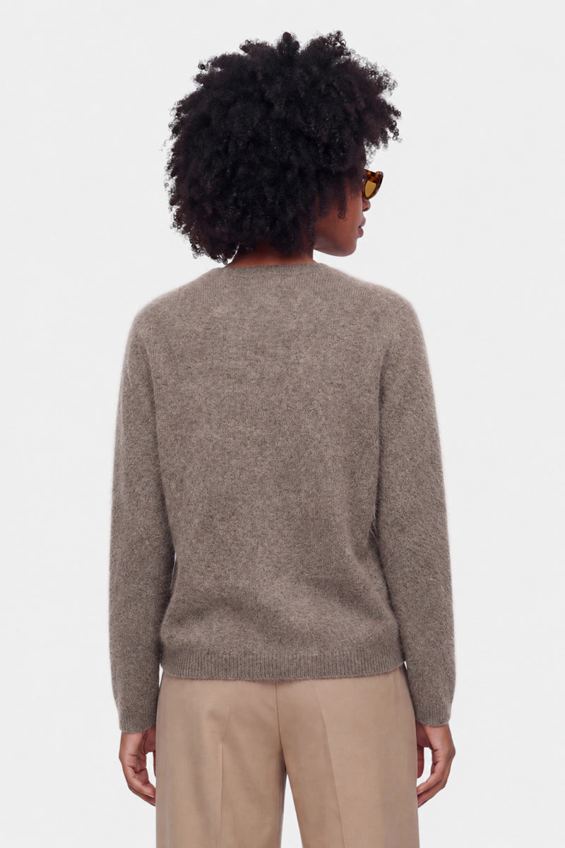 Eine Person mit kurzen, lockigen, dunklen Haaren, die eine Sonnenbrille trägt, einen locker sitzenden, flauschigen Pullover mit Rundhalsausschnitt in Taupe und eine beige Hose, steht mit dem Rücken zu einem schlichten weißen Hintergrund. Nur der obere Rücken und der Kopf sind vollständig zu sehen.
