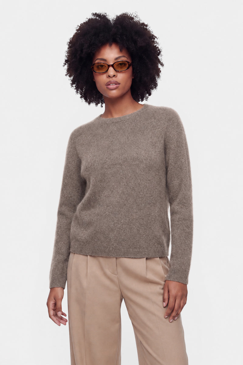 Eine Person mit lockigem Haar und brauner Sonnenbrille trägt einen flauschigen Pullover aus gebürsteter Wolle mit Rundhalsausschnitt und eine beige Hose und steht in einer entspannten Pose vor einem schlichten weißen Hintergrund.
