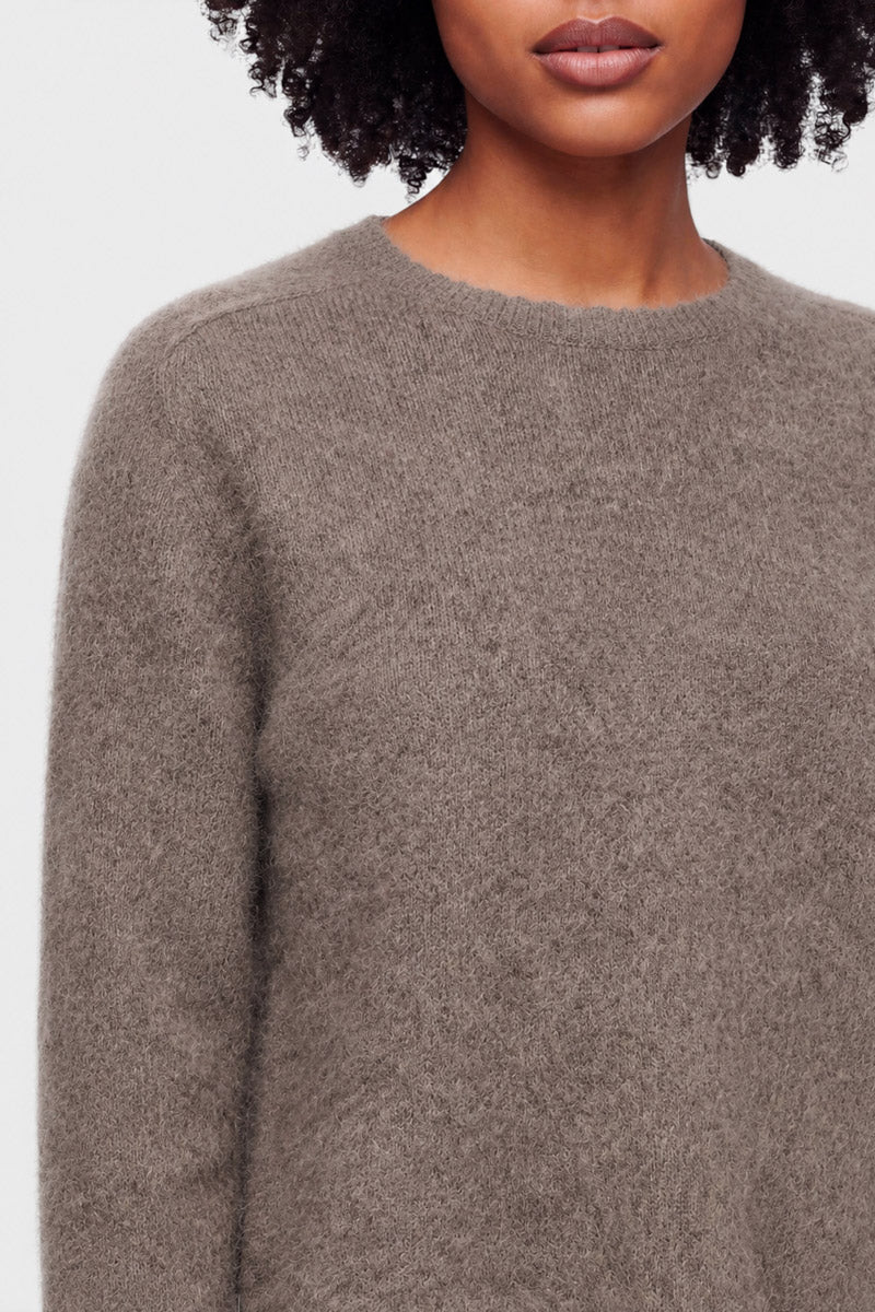 Eine Frau mit mittelbrauner Haut und lockigem Haar trägt einen flauschigen Pullover aus gebürsteter Wolle mit Rundhalsausschnitt in einem sanften Taupeton. Nur ihr Kopf (teilweise sichtbar, Lippen geschlossen) und ihr oberer Oberkörper sind vor einem schlichten, hellgrauen Hintergrund zu sehen. Es sind keine Accessoires oder Muster erkennbar.