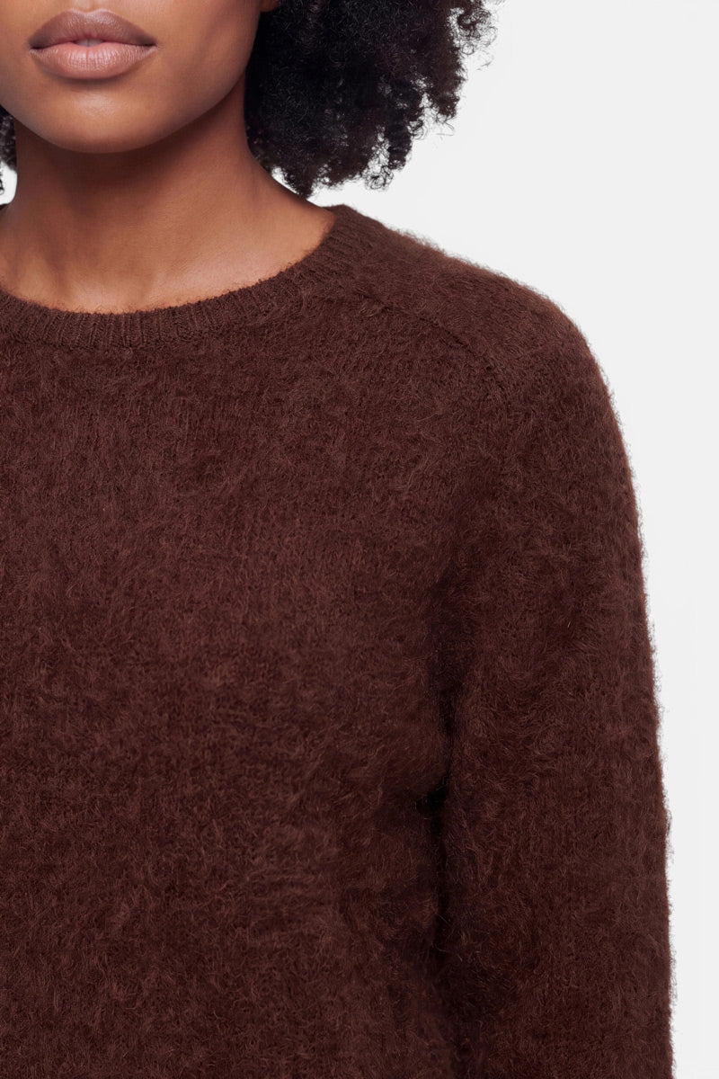 Eine Nahaufnahme einer Frau, die einen flauschigen Pullover aus strukturiertem, flauschigem, dunkelbraunem Stoff trägt. Zu sehen sind nur die untere Hälfte ihres Gesichts, ihre linke Schulter und ihr oberer Oberkörper. Sie hat mittelbraune Haut und dunkles, lockiges Haar; der Hintergrund ist schlicht weiß.