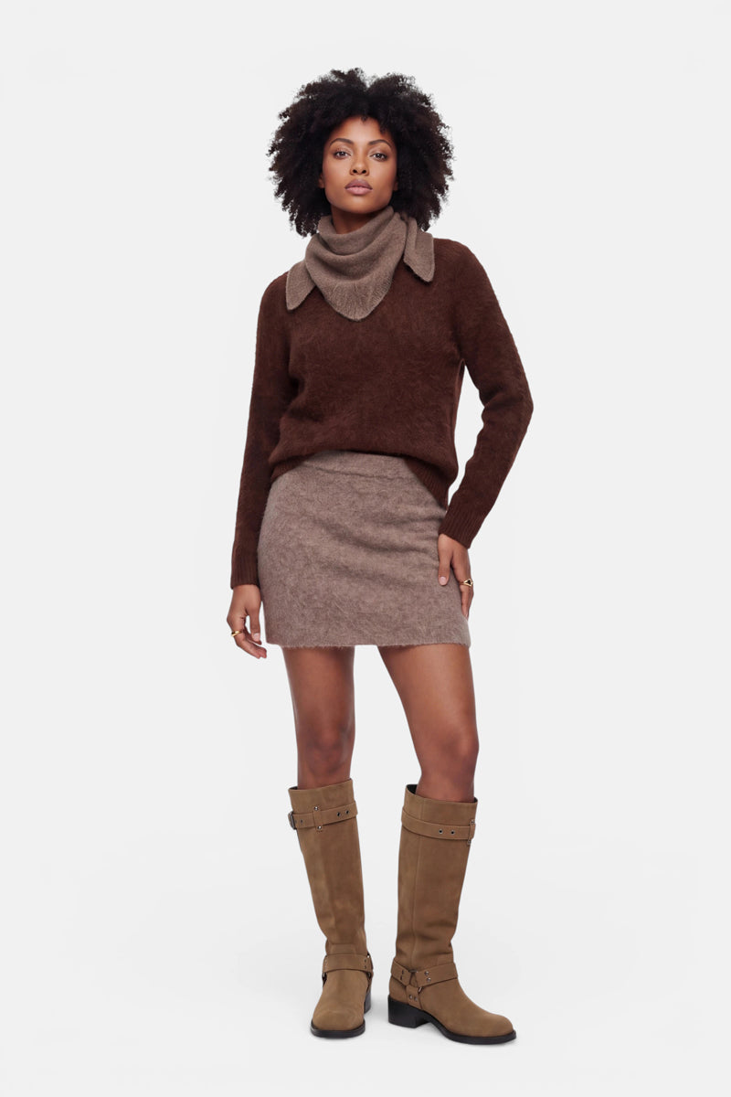 Eine Frau steht vor einem schlichten weißen Hintergrund und trägt einen braunen Pullover mit weitem Kragen sowie einen flauschigen Minirock aus gebürsteter Wolle mit elastischem Bund. Dazu trägt sie kniehohe, hellbraune Stiefel mit Schnallen. Ihr lockiges Haar und ihr neutraler Gesichtsausdruck unterstreichen ihren entspannten, ungezwungenen Stil, während ihre Arme ganz natürlich an den Seiten liegen.
