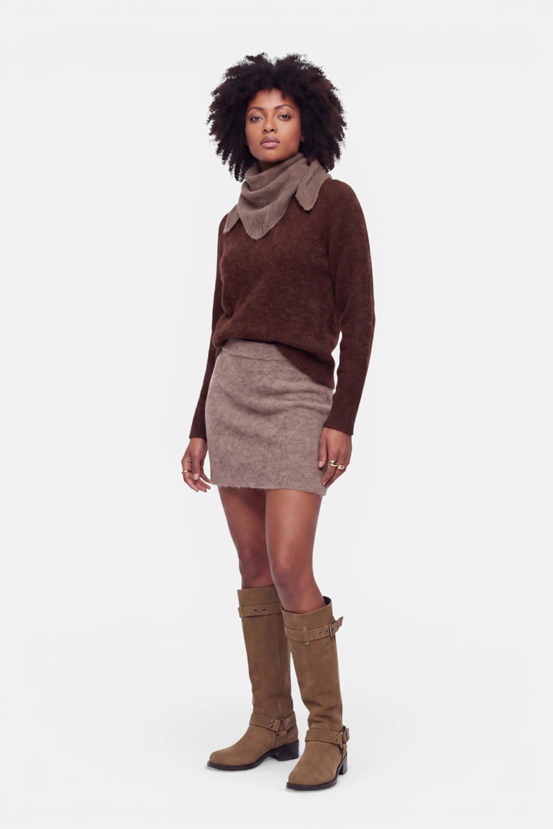 Eine Frau steht vor einem weißen Hintergrund und trägt einen dunkelbraunen Pullover mit großem Kragen, einen flauschigen Minirock aus gebürsteter Wolle mit elastischem Bund sowie kniehohe, hellbraune Stiefel mit Schnallen. Sie hat natürliches Afro-Haar, einen neutralen Gesichtsausdruck und lässt die Arme entspannt an den Seiten hängen.