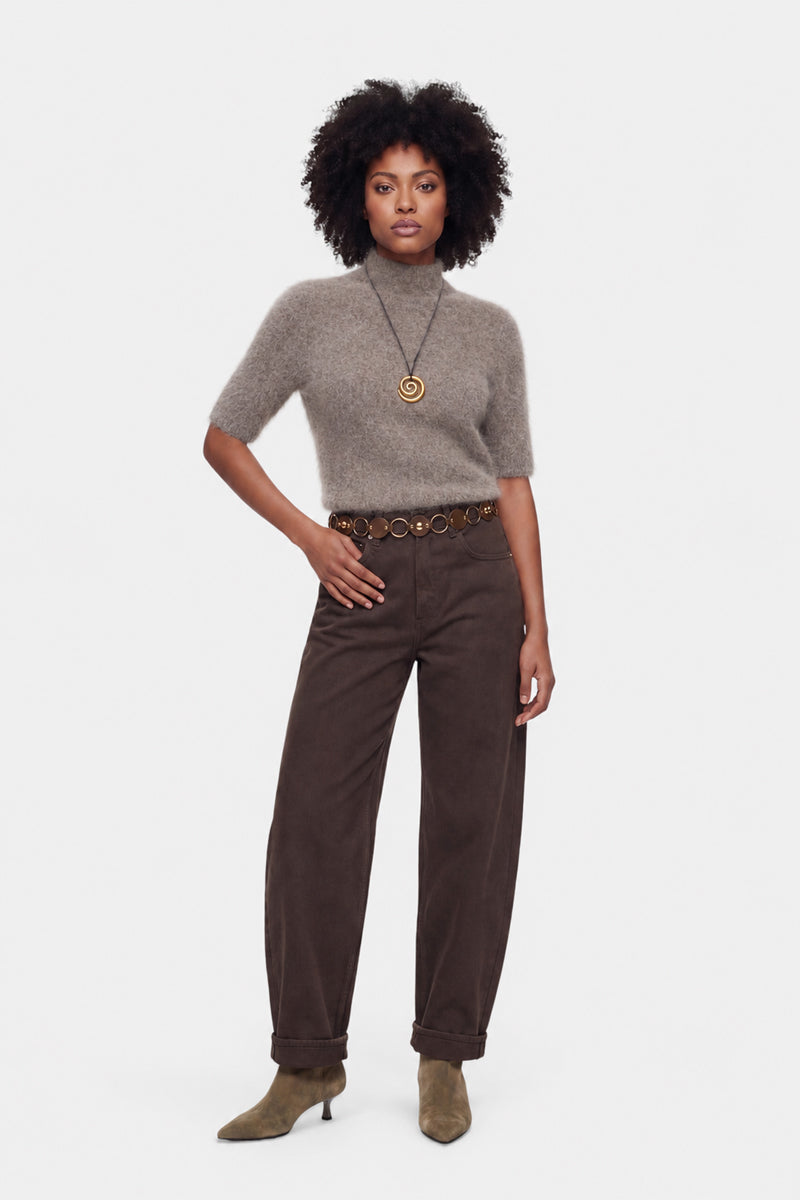 Eine Frau steht mit dem Blick nach vorne und trägt einen flauschigen Pullover – einen grauen, kurzärmeligen Pullover mit flauschiger Oberfläche –, eine braune, hoch taillierte Hose mit weitem Bein, olivfarbene Stiefeletten, eine Halskette mit rundem Anhänger sowie einen braunen Gürtel mit großen runden Akzenten. Vor einem schlichten Hintergrund posiert sie mit einer Hand in der Hüfte.