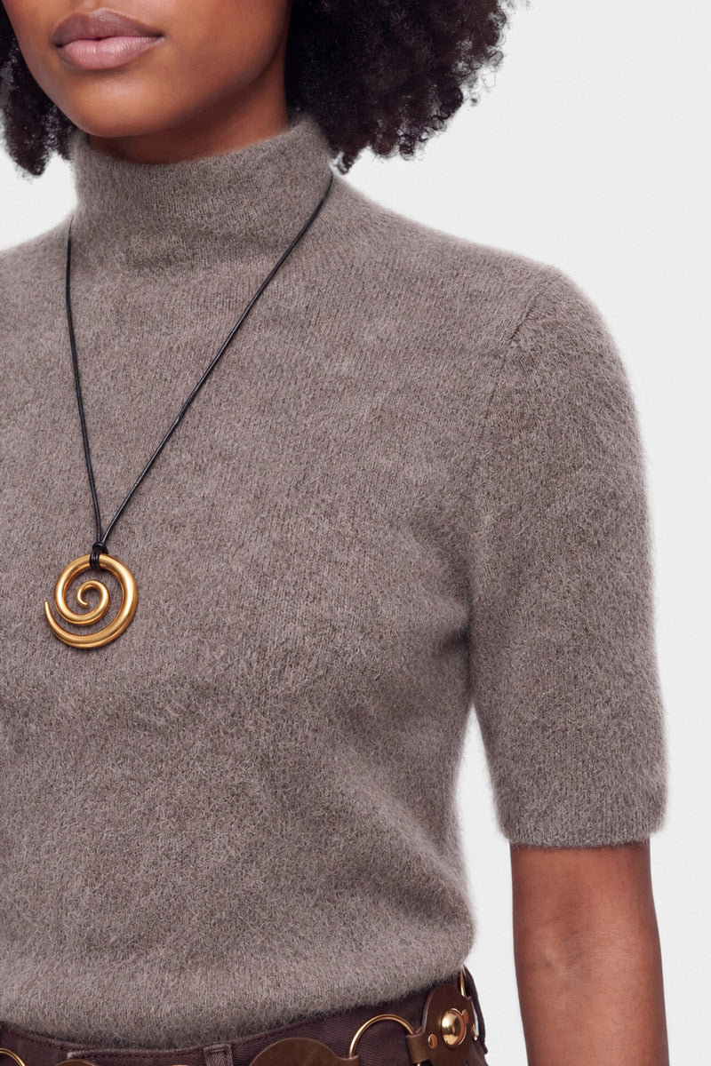 Eine Person trägt einen taupefarbenen Rollkragenpullover mit kurzen Ärmeln, eine Halskette mit spiralförmigem Anhänger und einen breiten braunen Gürtel. Zu sehen sind nur der untere Teil des Gesichts, der Hals und der obere Oberkörper; der Hintergrund ist einfarbig und hellgrau.