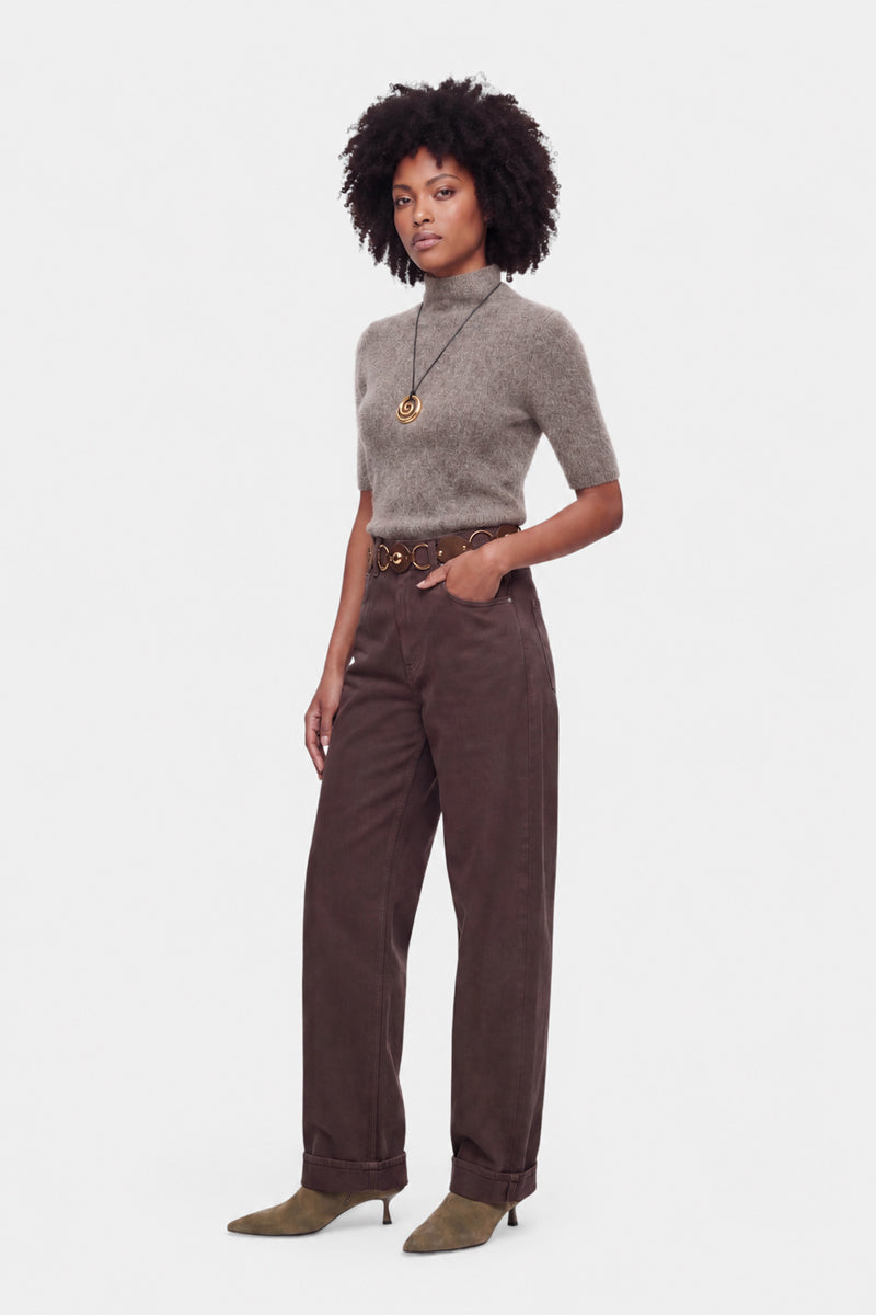Vor einem weißen Hintergrund steht eine Frau mit lockigem Haar. Sie trägt einen grauen, kurzärmeligen Rollkragenpullover, eine braune Hose mit hoher Taille und weitem Bein, olivgrüne Stiefel mit Absatz, einen schwarzen Gürtel mit goldener Schnalle und eine lange Halskette mit einem runden Anhänger.