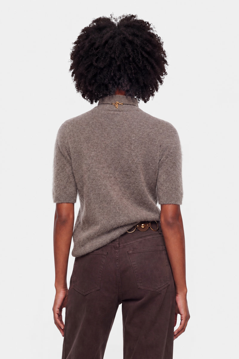 Eine Person steht mit dem Rücken zur Kamera und trägt einen kurzärmeligen, graubraunen Strickpullover aus gebürsteter Wolle sowie eine dunkelbraune Hose. Ihr lockiges Haar ist voluminös, und am Kragen des Pullovers ziert eine goldene Kette. An der Taille ist ein brauner Gürtel mit runden goldenen Akzenten zu sehen.