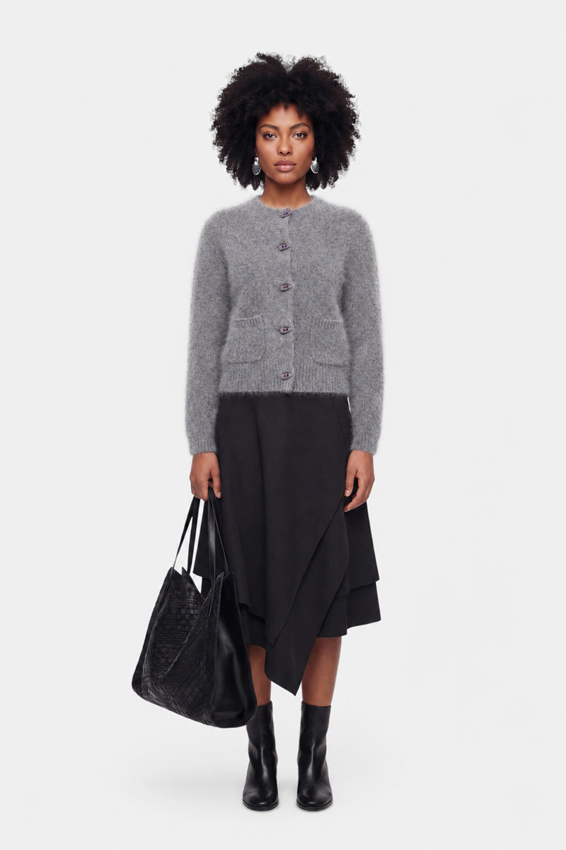 Eine Frau mit natürlich lockigem Haar trägt eine flauschige Strickjacke in Grau, einen mehrlagigen schwarzen Rock und schwarze Stiefeletten. Sie hält eine große schwarze, strukturierte Tragetasche in der Hand, steht vor einem schlichten weißen Hintergrund und blickt mit ausdruckslosem Gesichtsausdruck geradeaus.