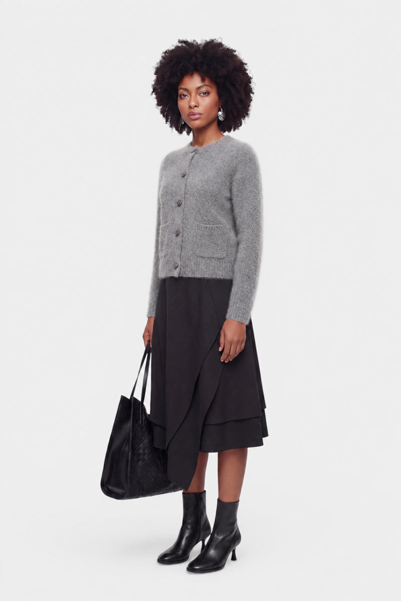 Eine Frau mit natürlich lockigem Haar trägt eine flauschige Strickjacke aus gebürsteter Wolle mit Rundhalsausschnitt, einen schwarzen, gestuften Midirock und schwarze Stiefeletten. Sie hält eine große, schwarze, geflochtene Tragetasche in der Hand und steht vor einem schlichten weißen Hintergrund, wobei sie mit neutralem Gesichtsausdruck nach vorne blickt.