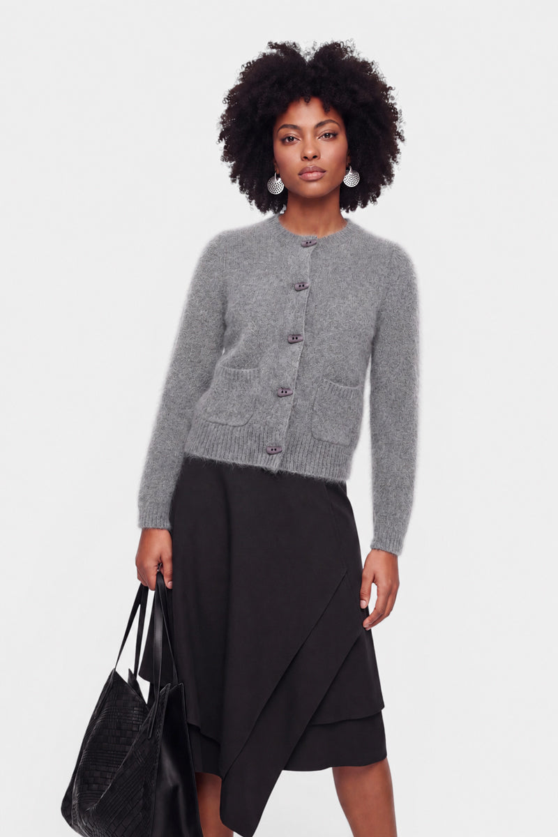 Eine Frau mit natürlich lockigem Haar trägt eine flauschige, graue Strickjacke mit Knöpfen, einen schwarzen Wickelrock und Ohrringe; in der Hand hält sie eine große, schwarze, strukturierte Tragetasche. Sie steht vor einem schlichten weißen Hintergrund und blickt selbstbewusst in die Kamera.