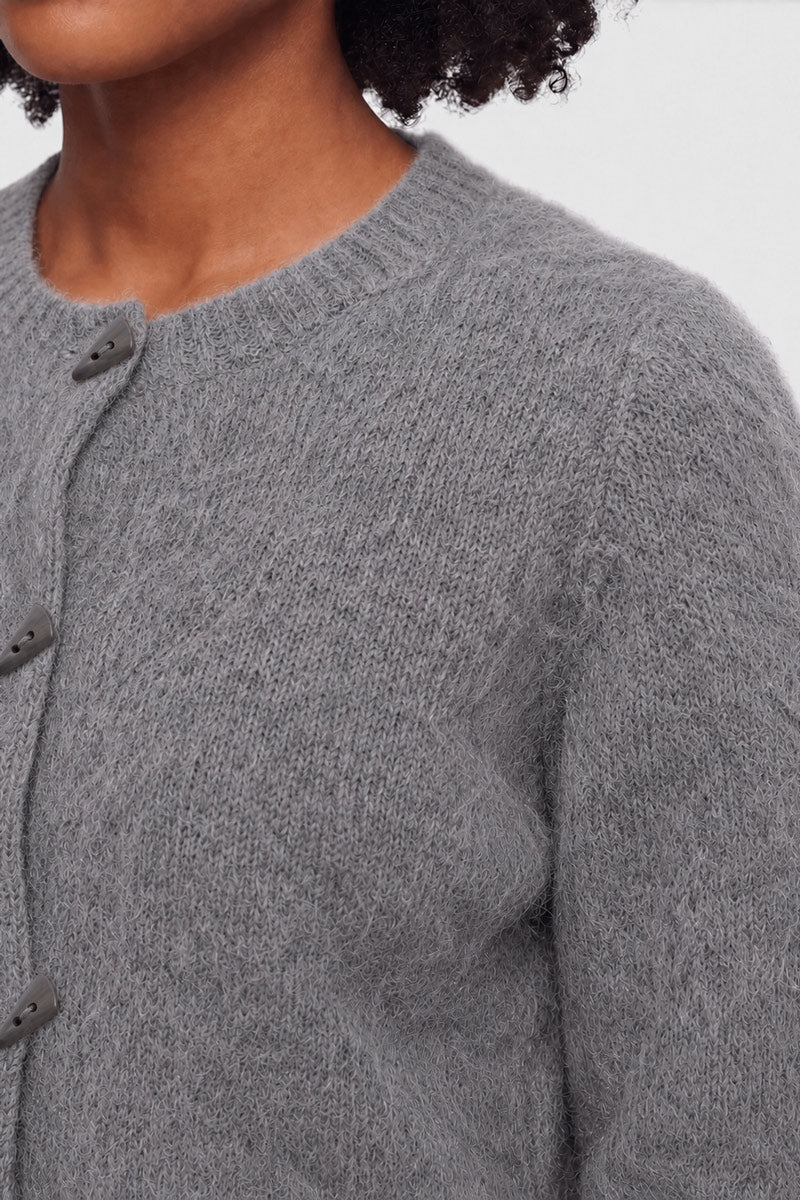 Nahaufnahme einer Person, die eine weiche, flauschige, graue Strickjacke mit Rundhalsausschnitt und Knopfleiste trägt. Zu sehen sind nur die untere Hälfte des Gesichts, der Hals und der obere Brustbereich. Der Hintergrund ist einfarbig und hell. Die Struktur des Pullovers ist deutlich zu erkennen.