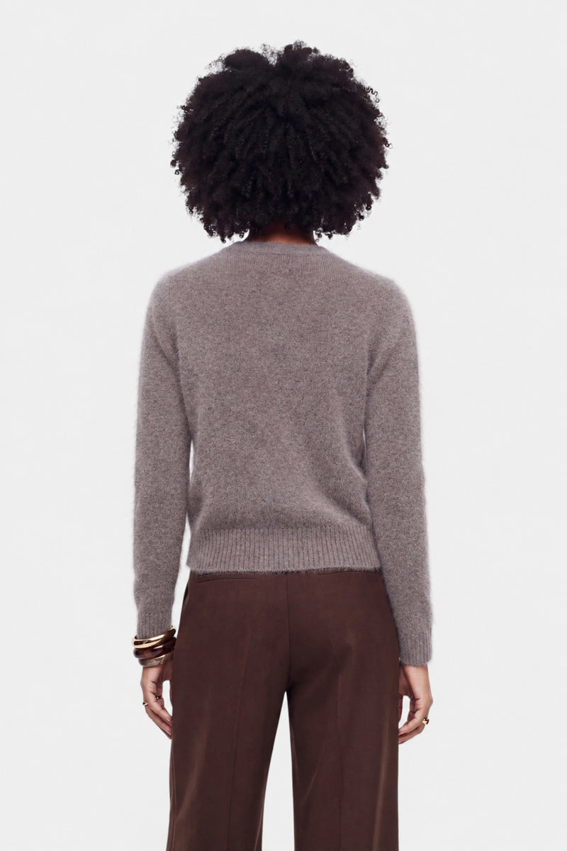 Eine Person steht mit dem Rücken zur Kamera und trägt eine flauschige Strickjacke mit Rundhalsausschnitt in zartem Hellgrau sowie eine braune Hose mit hoher Taille. Sie hat eine natürliche Afro-Frisur und trägt mehrere goldene Armreifen am linken Handgelenk. Der Hintergrund ist schlicht weiß.