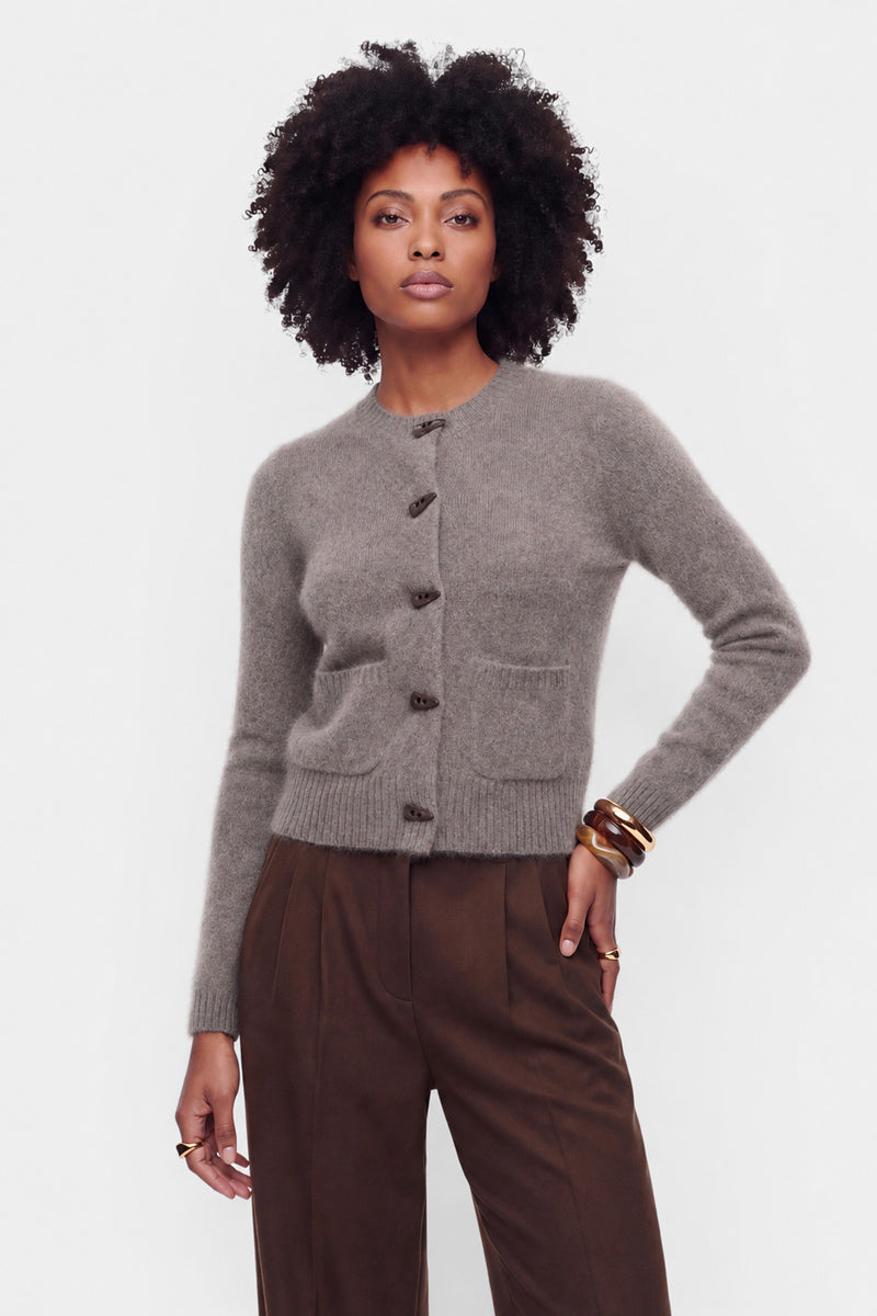 Eine Frau mit natürlich lockigem Haar trägt eine eng anliegende, geknöpfte Strickjacke aus gebürsteter Wolle mit Rundhalsausschnitt und eine braune Hose mit hoher Taille. Als Accessoires trägt sie goldene Armreifen an beiden Handgelenken und steht vor einem schlichten weißen Hintergrund, wobei sie selbstbewusst in die Kamera blickt.