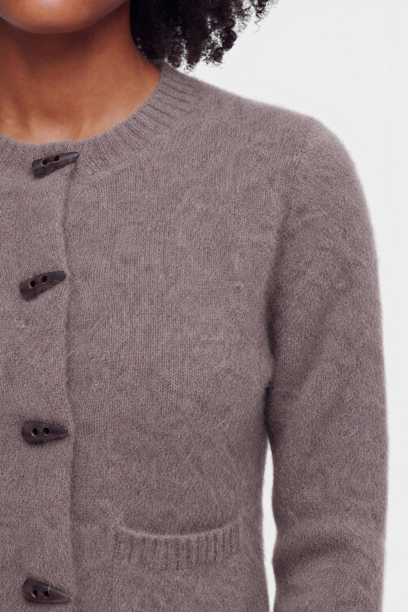 Eine Person, die eine flauschige Strickjacke in Hellbraun mit schwarzen Knöpfen und einer kleinen Brusttasche trägt, ist von den Schultern bis zur Brust zu sehen. Die Strickjacke hat einen klassischen Rundhalsausschnitt, und vor einem schlichten, hellen Hintergrund sind nur der untere Teil des Gesichts, der Hals und der obere Oberkörper zu sehen.