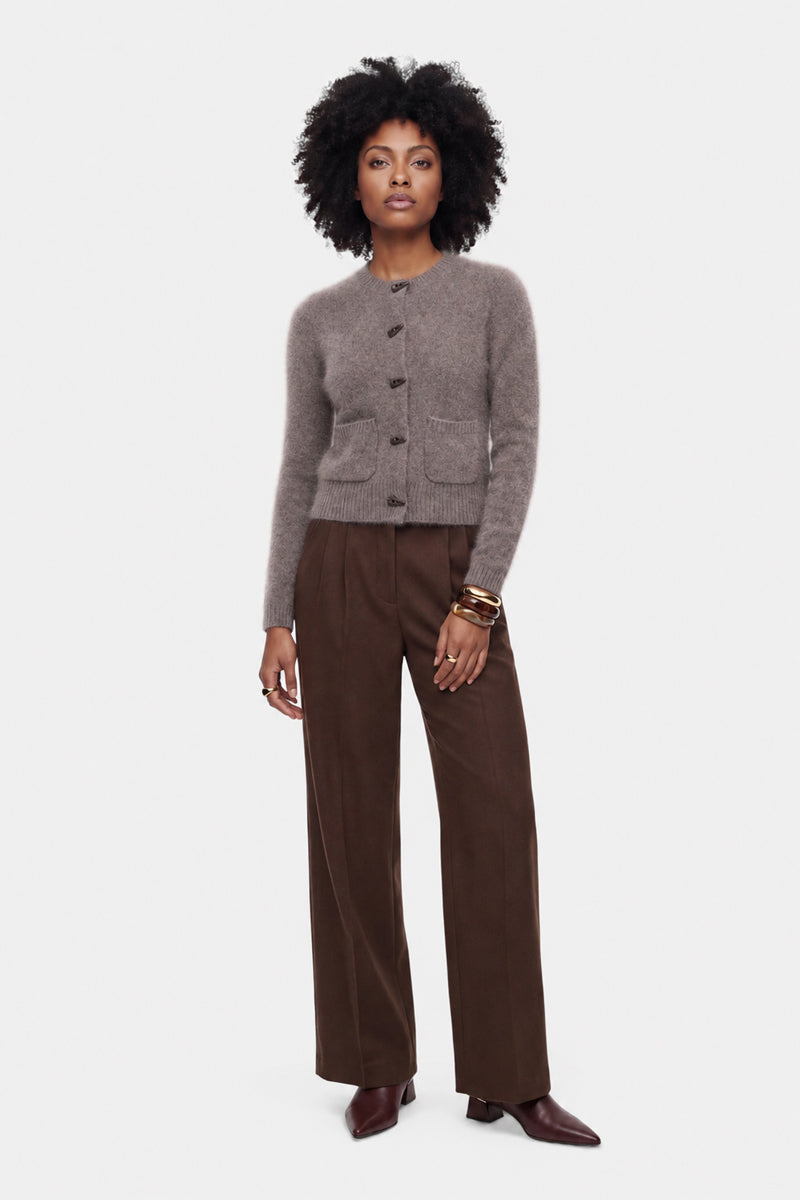 Eine Frau steht mit dem Blick nach vorne und trägt eine flauschige Strickjacke mit Rundhalsausschnitt und zwei Taschen, kombiniert mit weiten braunen Hosen und spitzen braunen Schuhen. Sie hat eine Afrofrisur, trägt goldene Creolen und mehrere goldene Armreifen und steht vor einem schlichten weißen Hintergrund.
