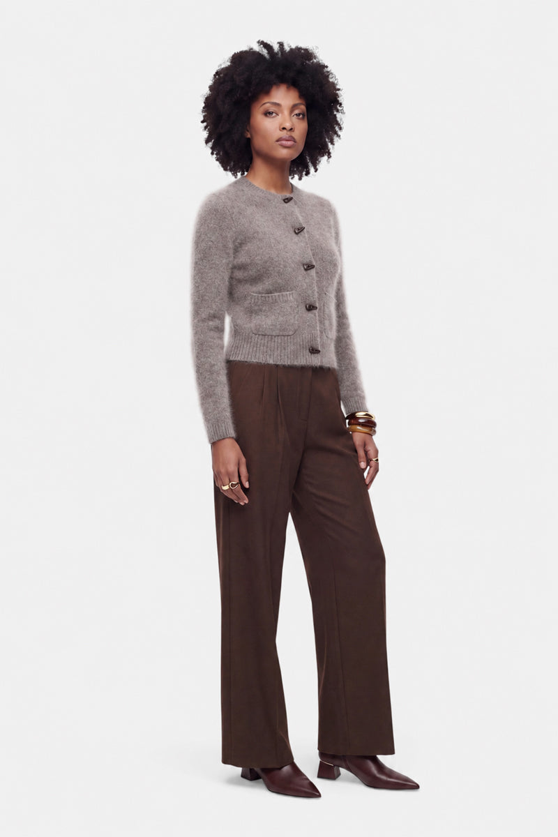 Vor einem weißen Hintergrund steht eine Frau, die eine flauschige Strickjacke aus gebürsteter Wolle mit Rundhalsausschnitt trägt, kombiniert mit einer braunen Hose mit weitem Bein und braunen spitzen Stiefeln. Sie hat natürlich lockiges Haar und trägt goldene Armbänder und Ringe, während sie selbstbewusst in die Kamera blickt.