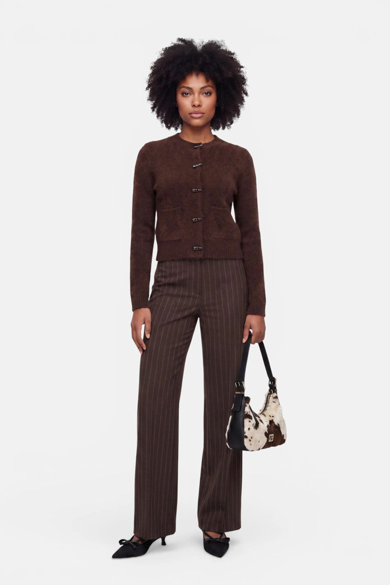 Eine Frau mit natürlichem Afro-Haar trägt eine braune Strickjacke aus gebürsteter Wolle, eine braune Nadelstreifenhose und schwarze Pumps mit spitzer Zehenpartie. In der Hand hält sie eine schwarz-weiße Handtasche mit Kuhmuster. Der Hintergrund ist schlicht weiß, was ihr stilvolles, aufeinander abgestimmtes Outfit und ihre selbstbewusste Haltung unterstreicht.