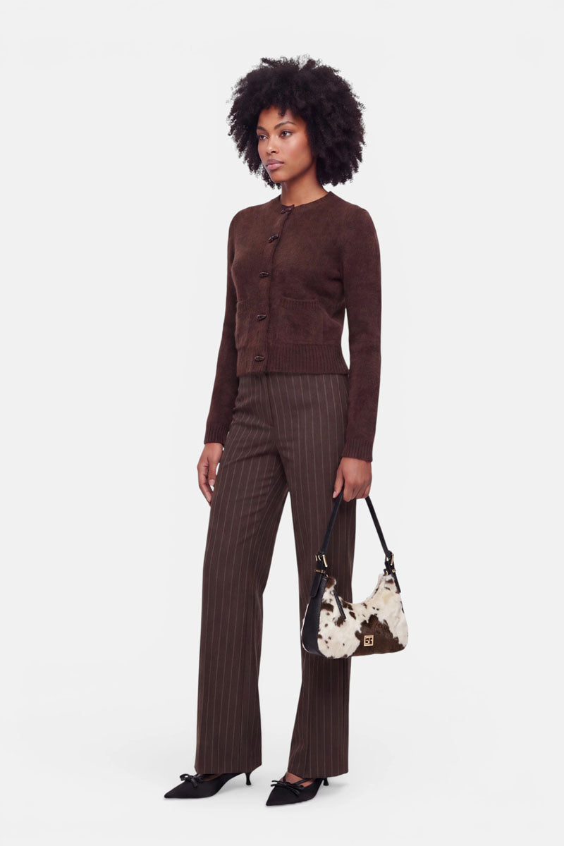 Vor einem weißen Hintergrund steht eine Frau, die eine dunkelbraune Strickjacke aus gebürsteter Wolle, eine braune Nadelstreifenhose und schwarze spitze Pumps trägt und eine kleine schwarz-weiße, pelzbesetzte Handtasche in der Hand hält. Sie hat schulterlanges lockiges Haar und einen neutralen Gesichtsausdruck.