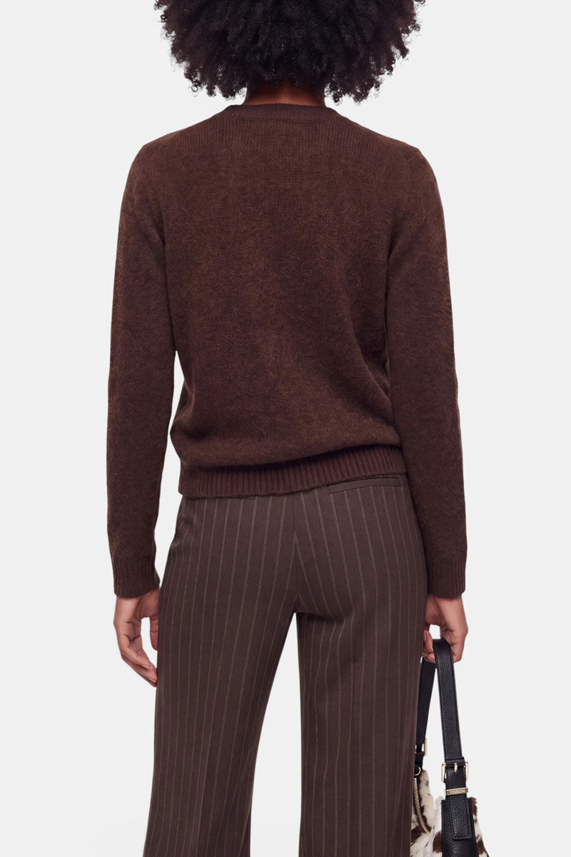 Eine Person steht mit dem Rücken zur Kamera und trägt eine dunkelbraune Strickjacke aus gebürsteter Wolle sowie eine braune Nadelstreifenhose. In der linken Hand hält sie eine Handtasche. Das lockige Haar der Person ist zu sehen, der Hintergrund ist schlicht weiß.