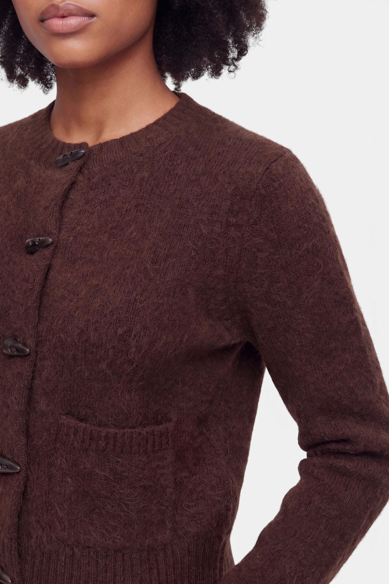 Eine Frau mit lockigem Haar trägt eine flauschige Strickjacke in einem satten Braunton mit strukturierter Oberfläche, Rundhalsausschnitt, Knebelknöpfen und einer aufgesetzten Brusttasche. Das Foto ist vom Hals bis zur Mitte des Oberkörpers beschnitten und zeigt die linke Seite ihres Körpers vor einem einfarbigen Hintergrund.
