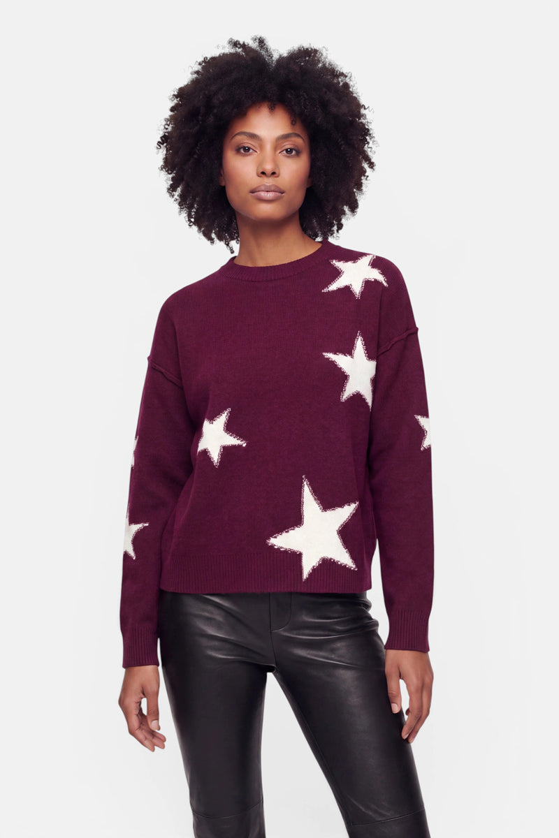 Eine Frau mit lockigem Haar trägt einen kastenförmigen Pullover – einen burgunderroten Pullover, der mit großen weißen Sternen im Sternen-Intarsia-Stil verziert ist – sowie eine schwarze Lederhose. Sie steht mit neutralem Gesichtsausdruck vor einem schlichten weißen Hintergrund und blickt nach vorne.