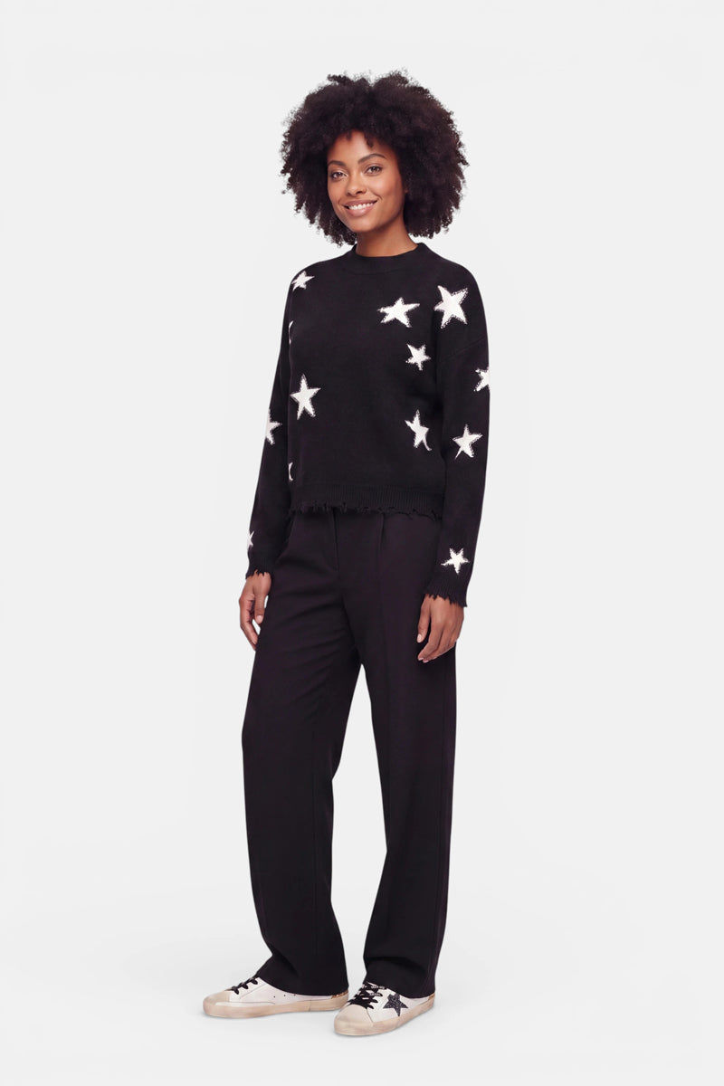 Eine Person mit lockigem Haar lächelt. Sie trägt einen Merino-Pullover mit weißer Sternen-Intarsia, eine weite schwarze Hose und weiße Turnschuhe mit schwarzen Streifen. Sie steht vor einem schlichten weißen Hintergrund.