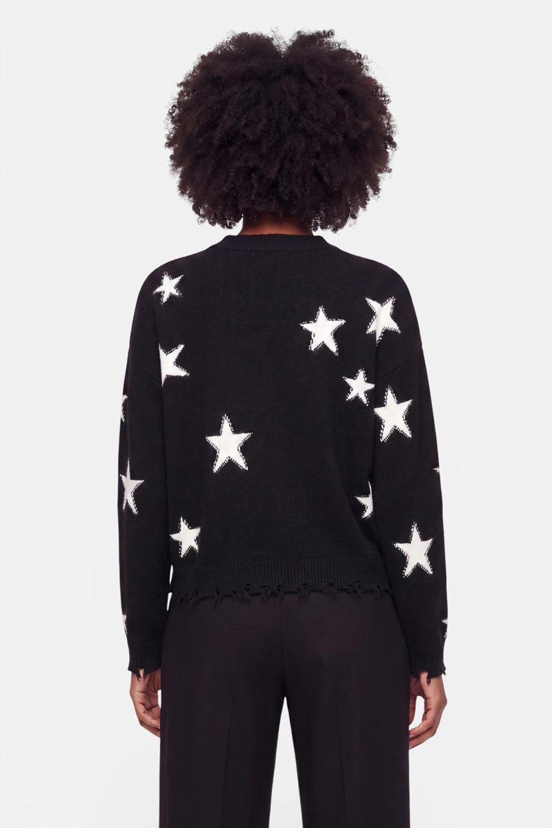 Eine Person mit lockigem Haar steht mit dem Rücken zur Kamera und trägt einen schwarzen Damenpullover mit großem weißem Sternen-Intarsia-Muster. Der Merino-Pullover hat eine lockere Passform und einen leicht ausgefransten Saum und wird vor einem schlichten weißen Hintergrund mit einer weiten schwarzen Hose kombiniert.