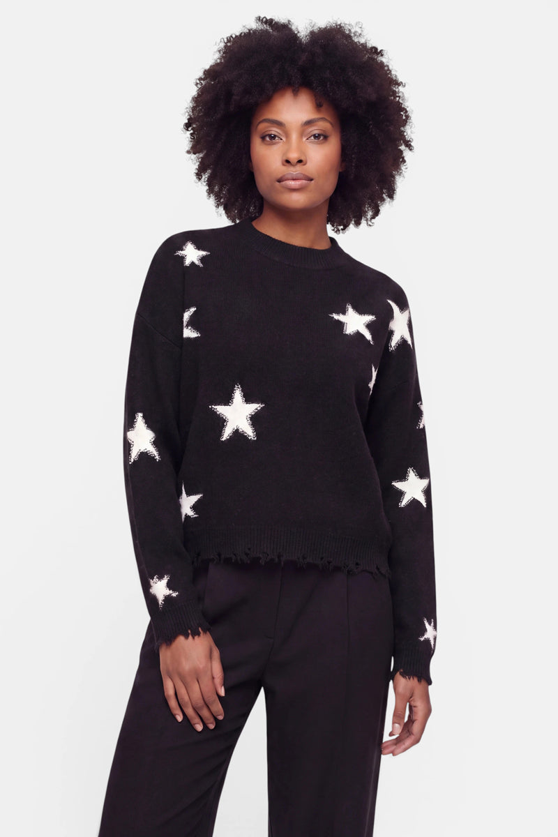 Eine Frau mit lockigem Haar trägt einen schwarzen Merino-Pullover mit großen weißen Sternen-Intarsienmustern. Der Pullover hat gerippte Bündchen und einen gerippten Saum, und sie kombiniert ihn mit einer schwarzen Hose. Sie steht vor einem schlichten weißen Hintergrund und blickt selbstbewusst in die Kamera.