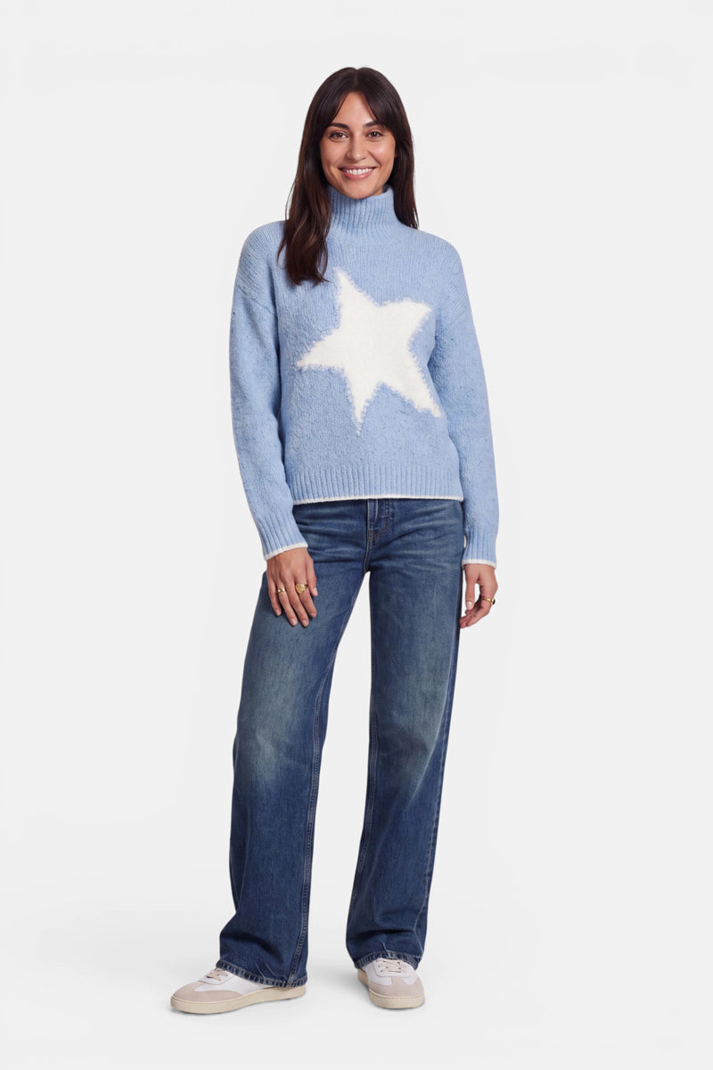 Eine Frau mit glattem, dunklem Haar trägt einen hellblauen Grobstrickpullover im Oversize-Design mit einem großen weißen Stern, eine blaue Jeans und weiße Sneaker. Sie steht vor einem schlichten weißen Hintergrund und lächelt.