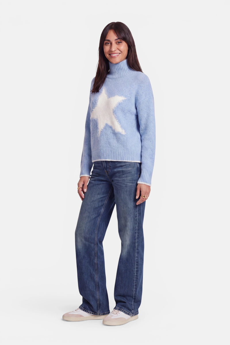 Eine Frau mit langen braunen Haaren lächelt. Sie trägt einen hellblauen Grobstrickpullover mit einem großen weißen Stern auf der Vorderseite, der sich durch ein Oversize-Design und einen kuscheligen Stehkragen auszeichnet. Dazu trägt sie eine blaue Jeans und weiße Schuhe vor einem schlichten weißen Hintergrund.