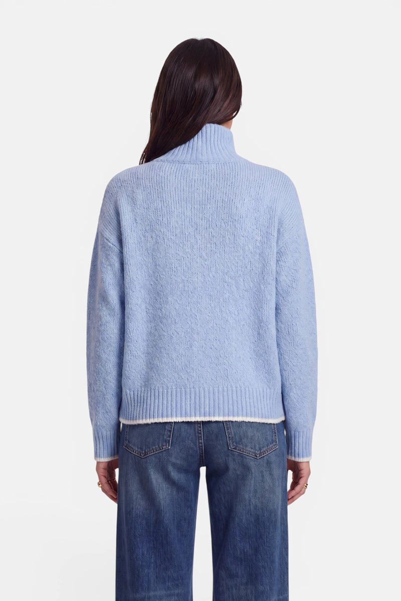 Eine Person steht mit dem Rücken zur Kamera und trägt einen hellblauen Grobstrickpullover im Oversize-Design sowie eine klassische Bluejeans. Der Pullover hat einen gerippten Saum, gerippte Ärmelbündchen und einen Stehkragen. Das lange, dunkle Haar der Person fällt zur Seite. Der Hintergrund ist schlicht weiß.