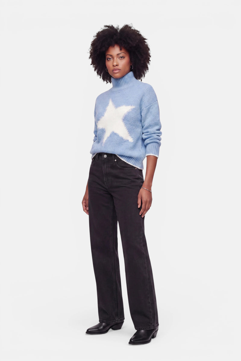 Vor einem weißen Hintergrund steht eine Frau, die einen hellblauen Grobstrickpullover mit einem großen weißen Stern auf der Vorderseite und einem kuscheligen Stehkragen trägt. Der Oversize-Pullover lässt sich mühelos mit einer schwarzen Hose mit weitem Bein und schwarzen Schuhen kombinieren. Sie hat dunkles, lockiges Haar und einen neutralen Gesichtsausdruck, die Arme entspannt an den Seiten.