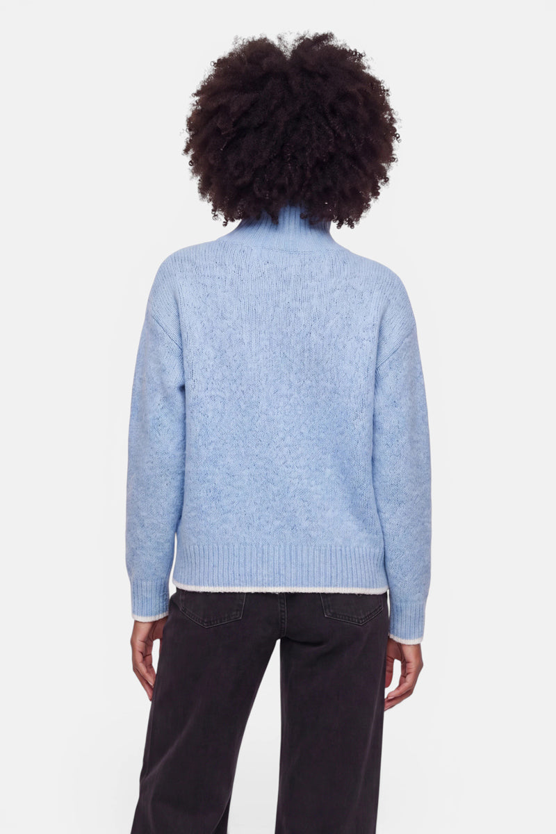 Eine Person mit lockigem Haar, die mit dem Rücken zur Kamera steht und einen hellblauen Grobstrickpullover mit Stehkragen sowie eine dunkle Hose trägt. Der Hintergrund ist schlicht weiß, und die Hände ruhen locker an den Seiten. Der Pullover wirkt weich und hat gerippte Bündchen an Ärmeln und Saum in einem Oversize-Design. Das Haar ist voluminös und natürlich.