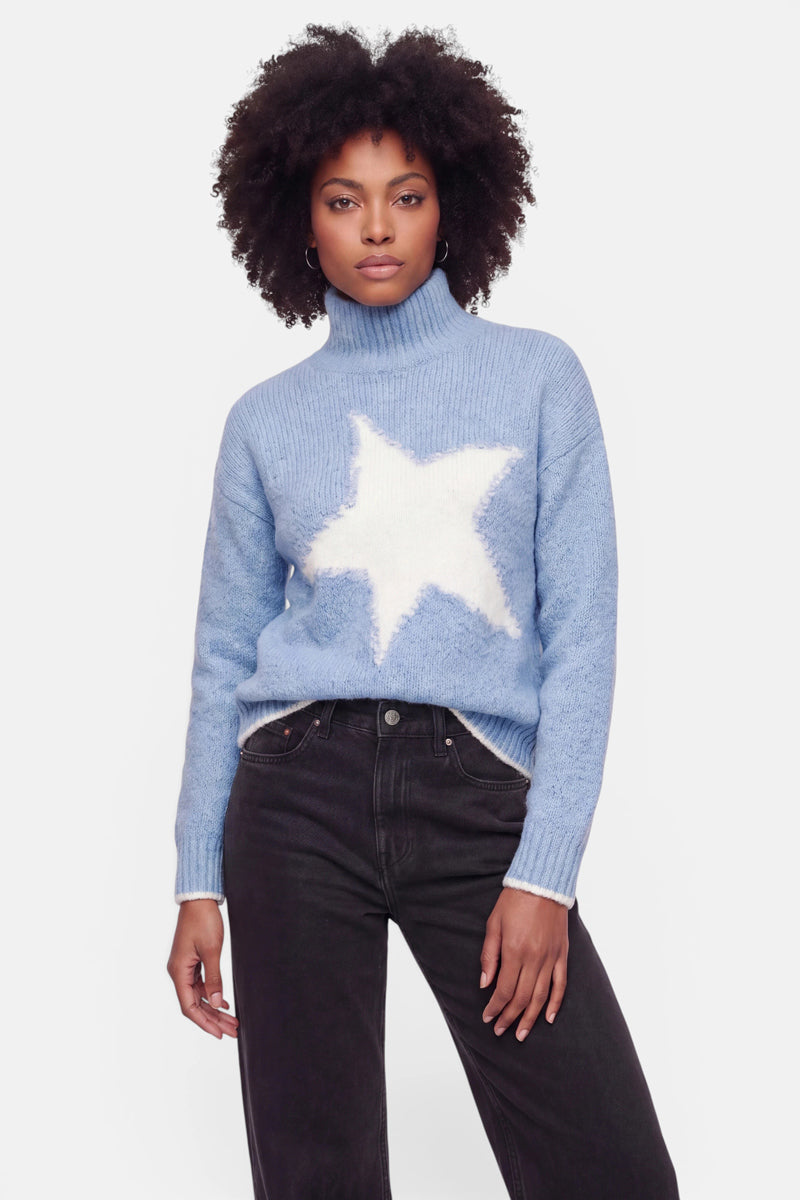 Eine Frau mit lockigem Haar trägt einen hellblauen Grobstrickpullover mit Stehkragen, auf dessen Vorderseite ein großer weißer Stern zu sehen ist, sowie eine schwarze Hose. Sie steht mit dem Blick nach vorne gerichtet, einen Arm entspannt an der Seite, und einem neutralen Gesichtsausdruck vor einem schlichten weißen Hintergrund.