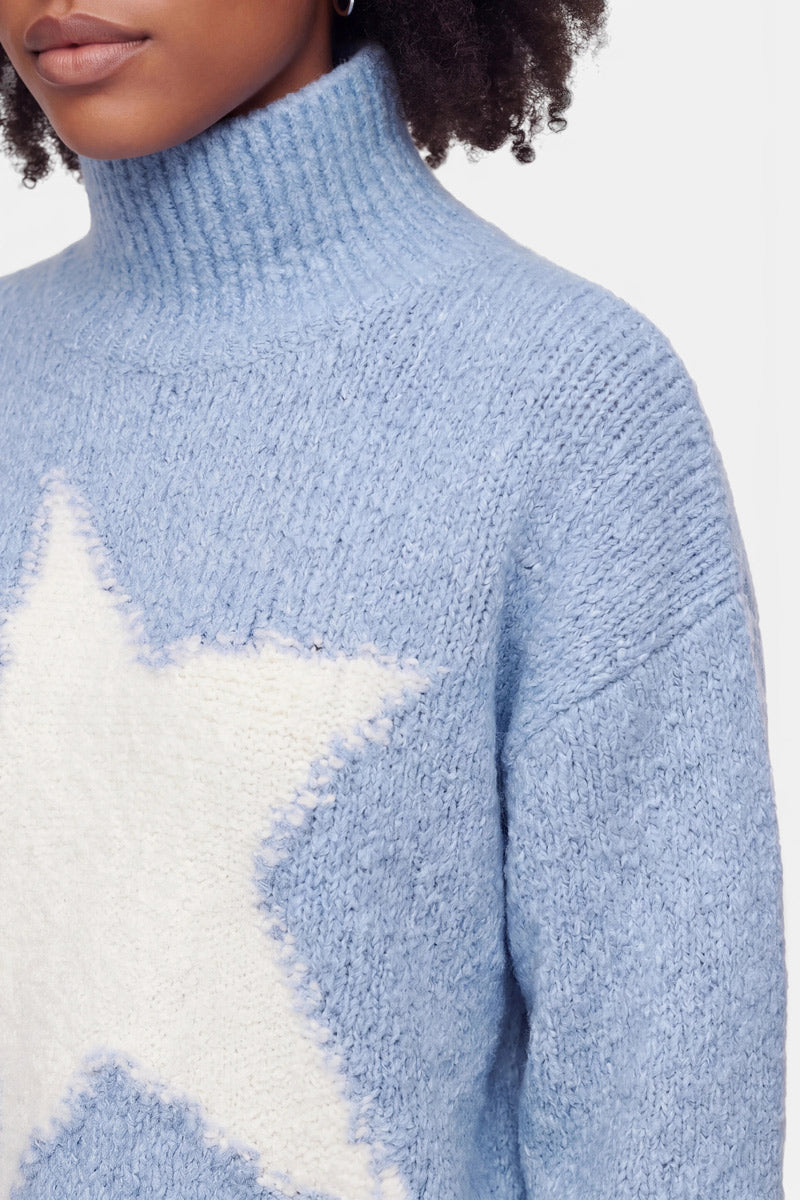 Eine Person, die einen hellblauen Rollkragenpullover im Grobstrick-Stil mit einem großen weißen Stern auf der Brust trägt. Zu sehen sind nur der untere Teil des Gesichts, der Hals und der obere Oberkörper. Die Person hat lockiges Haar und steht vor einem einfarbigen Hintergrund.