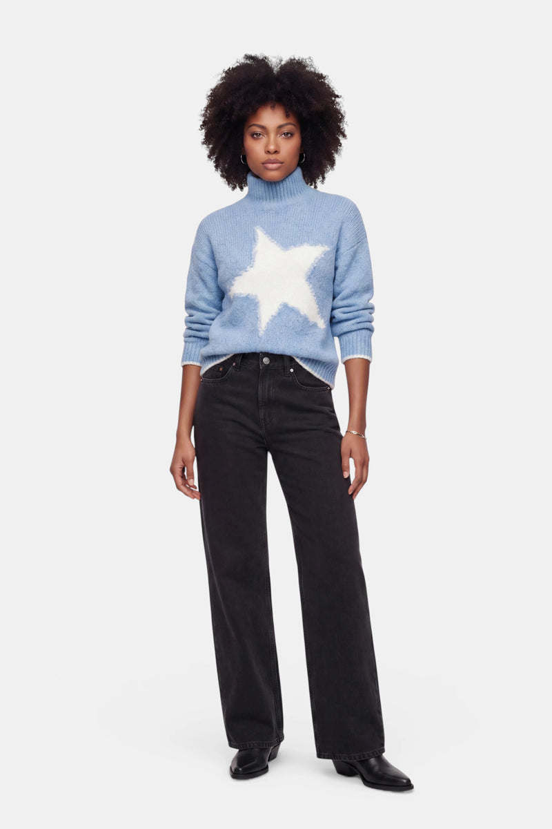 Eine Frau steht mit dem Blick nach vorne und trägt einen hellblauen Grobstrickpullover mit Stehkragen und einem großen weißen Stern auf der Vorderseite, dazu eine schwarze Jeans mit hoher Taille und weitem Bein sowie schwarze Schuhe. Sie hat natürlich lockiges Haar und einen neutralen Gesichtsausdruck. Der Pullover besticht durch sein Oversize-Design, das für zusätzlichen Komfort und Stil sorgt. Der Hintergrund ist schlicht weiß.