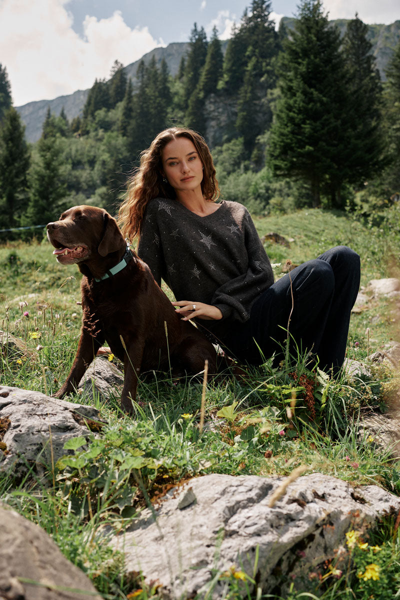 Eine Frau mit langen, welligen Haaren, die einen dunklen Pullover aus Alpaka-Wolle mit V-Ausschnitt trägt, sitzt neben einem braunen Hund im Gras. Hinter ihnen sind Felsen, Wildblumen, grüne Kiefern und ferne Berge unter einem teilweise bewölkten Himmel zu sehen. Dezente Strassstein-Sterne verleihen ihrem kuscheligen Pullover einen Hauch von Glanz.