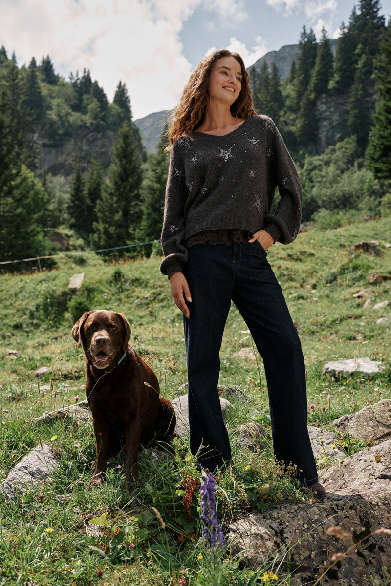 Eine lächelnde Frau in einem dunklen Pullover aus Alpaka-Wolle mit Sternenmuster, V-Ausschnitt und Jeans steht auf einer Wiese neben einem braunen Hund. Sie befinden sich im Freien in einer grünen Berglandschaft mit Bäumen, Felsen und Blumen unter einem teilweise bewölkten Himmel.