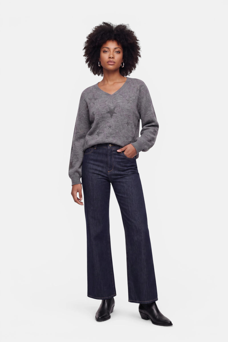 Eine Frau mit Afro-Frisur trägt einen locker sitzenden, grauen Pullover aus Alpaka-Wolle mit V-Ausschnitt und dezenten Strass-Sternmotiven, dazu dunkle, weit geschnittene Jeans und schwarze Stiefeletten. Sie steht selbstbewusst vor einem schlichten weißen Hintergrund, eine Hand in der Jeanstasche.