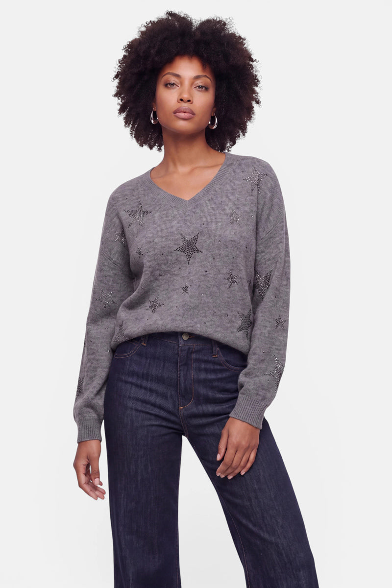 Eine Frau mit natürlich lockigem Haar trägt einen grauen Pullover aus Alpaka-Wolle mit V-Ausschnitt, der dezent mit zarten Sternmustern und Strasssteinen verziert ist, dazu eine dunkle Jeans mit hoher Taille. Sie steht vor einem schlichten weißen Hintergrund und blickt mit entspannter Haltung selbstbewusst in die Kamera.
