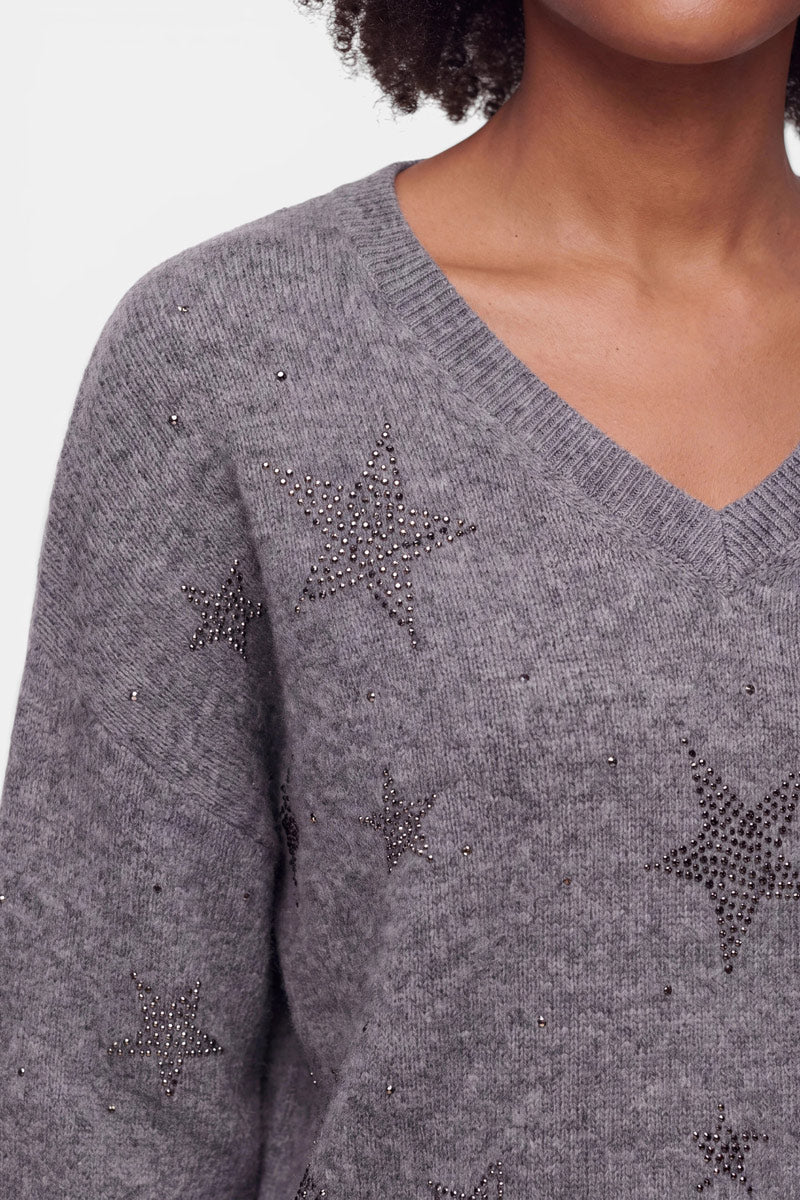 Eine Person, die einen grauen Strickpullover aus Alpaka-Wolle mit V-Ausschnitt trägt. Der Pullover ist mit funkelnden Strass-Sternenmotiven verziert, die über den Stoff verteilt sind. Nur der untere Teil des Gesichts und die Schulter sind zu sehen; der Hintergrund ist einfarbig weiß.