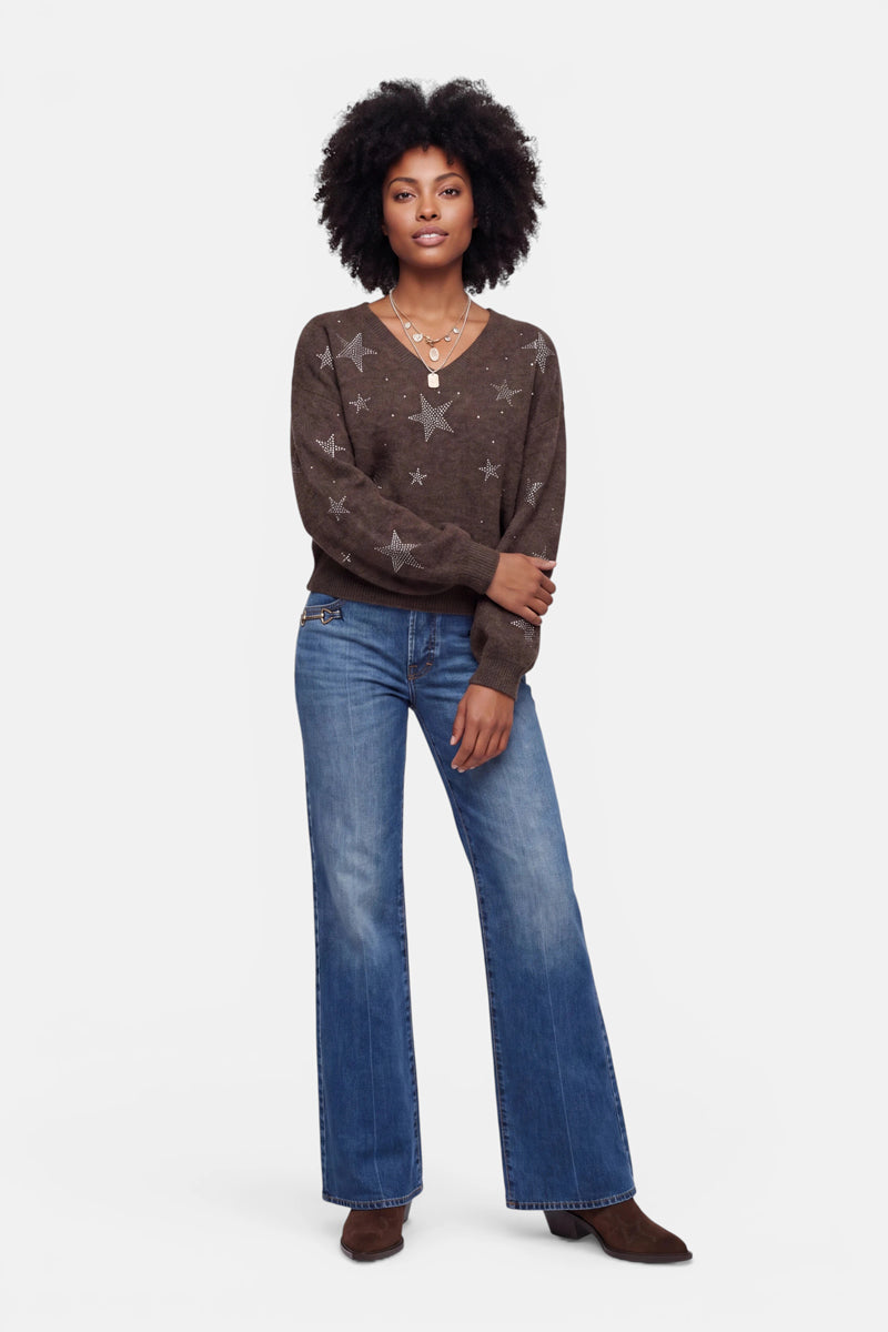 Eine Frau steht mit dem Blick nach vorne und trägt einen dunkelbraunen Pullover aus Alpaka-Wolle mit Sternmotiven, eine blaue Jeans mit weitem Bein, braune Schuhe und mehrere Halsketten übereinander. Sie hat dunkles, lockiges Haar und einen entspannten Gesichtsausdruck; ihre rechte Hand ruht auf ihrem linken Arm. Weißer Hintergrund.