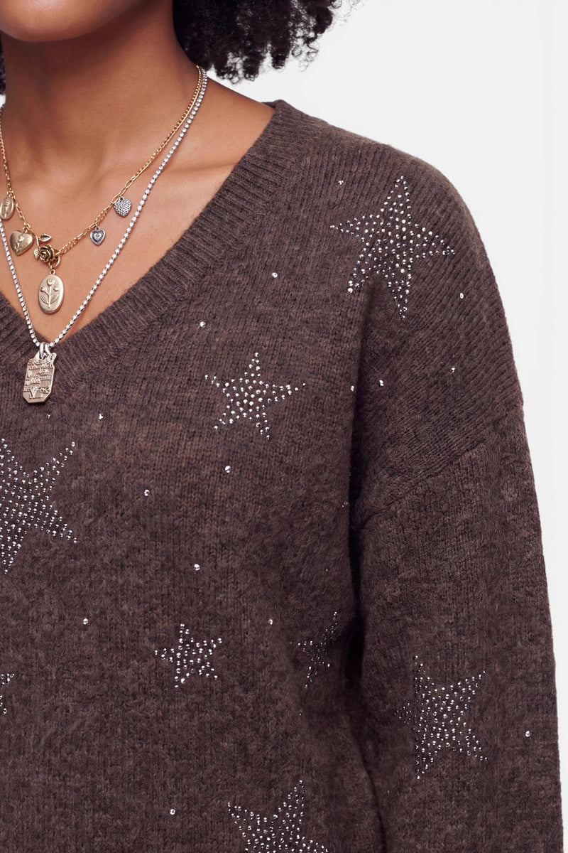 Eine Person, die mehrere goldene Halsketten trägt und einen braunen Pullover aus Alpaka-Wolle mit Strassstein-Sternen. Zu sehen sind nur der untere Teil des Gesichts, der Hals und der obere Oberkörper. Der Pullover hat einen V-Ausschnitt und eine weiche Struktur. Der Hintergrund ist einfarbig und hell.