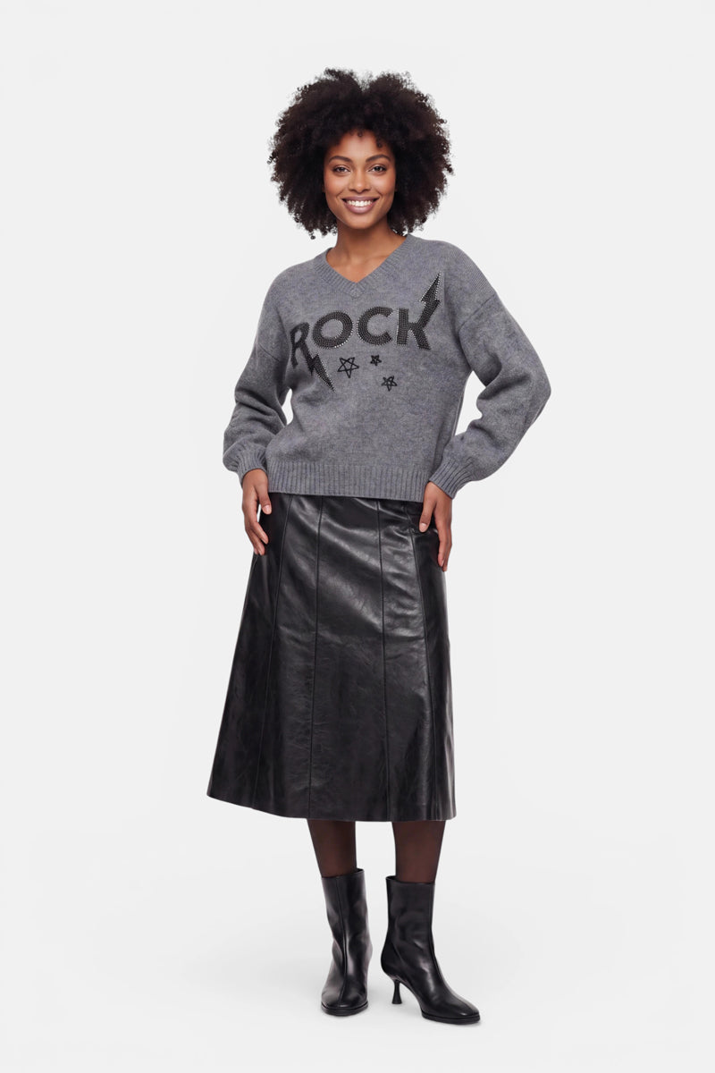 Eine lächelnde Frau trägt einen grauen Pullover aus Merinowolle mit schwarzen Sternen und dezenten Strassverzierungen, dazu einen schwarzen Midi-Lederrock mit eingenähten Einsätzen und schwarze Stiefeletten mit Absatz. Sie steht mit den Händen in den Hüften vor einem schlichten weißen Hintergrund und blickt nach vorne.