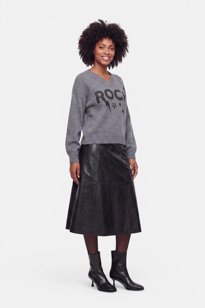 Eine Frau mit lockigem Haar trägt einen grauen Pullover aus Merinowolle mit V-Ausschnitt und Strassverzierung mit Sternen, einen schwarzen, plissierten Midi-Lederrock und schwarze Stiefeletten mit Absatz. Sie steht lächelnd vor einem schlichten weißen Hintergrund.