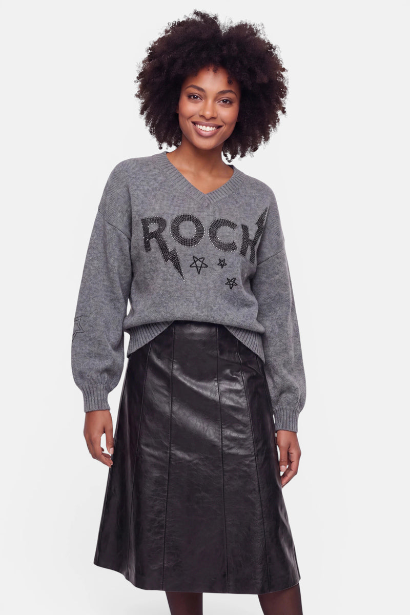 Eine Frau lächelt. Sie trägt einen locker sitzenden grauen Pullover aus Merinowolle mit „ROCK“- und Sternmotiven sowie dezenten Strassverzierungen, dazu einen schwarzen A-Linien-Rock aus Kunstleder. Sie hat mittellanges lockiges Haar und steht vor einem schlichten weißen Hintergrund, den Blick nach vorne gerichtet, die Arme entspannt an den Seiten.