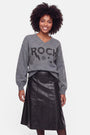 Eine Frau lächelt. Sie trägt einen locker sitzenden grauen Pullover aus Merinowolle mit „ROCK“- und Sternmotiven sowie dezenten Strassverzierungen, dazu einen schwarzen A-Linien-Rock aus Kunstleder. Sie hat mittellanges lockiges Haar und steht vor einem schlichten weißen Hintergrund, den Blick nach vorne gerichtet, die Arme entspannt an den Seiten.