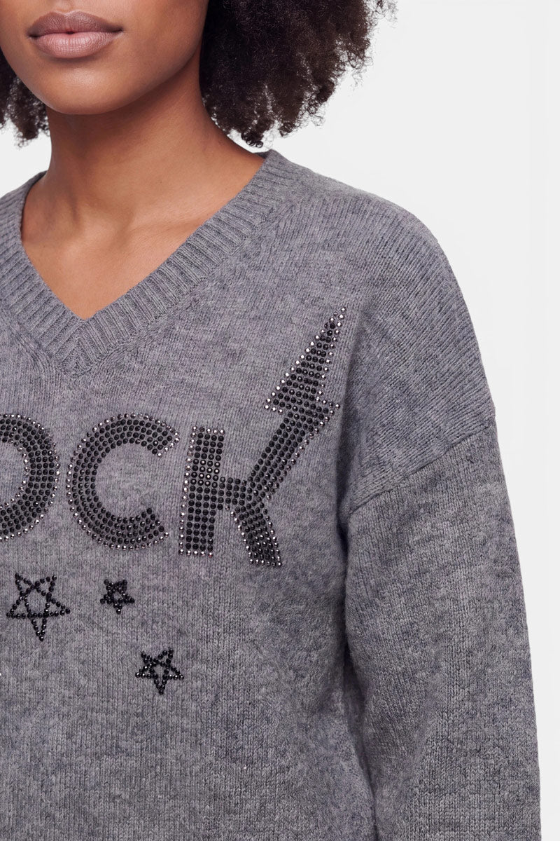 Eine Frau, die einen grauen Pullover aus Merinowolle mit V-Ausschnitt und Strassverzierung trägt, auf dem das Wort „ROCK“ zu lesen ist und der mit Sternmotiven verziert ist. Zu sehen sind nur ihr unteres Gesicht, ihr Hals und ihr oberer Oberkörper. Sie hat dunkles, lockiges Haar und hellbraune Haut. Der Hintergrund ist einfarbig und hell.