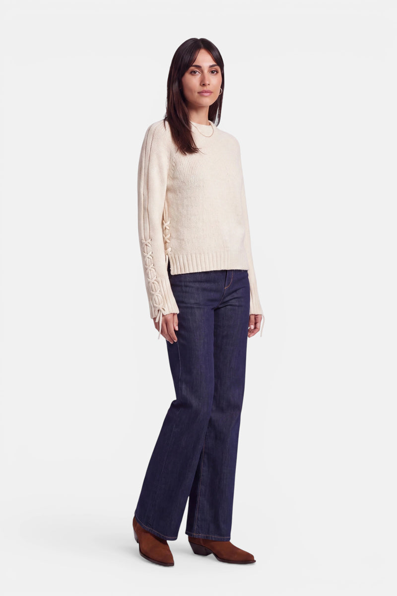Eine Frau mit glattem, dunklem Haar trägt einen cremefarbenen Pullover aus Alpaka-Wolle mit Raglanärmeln und eleganten Schnürdetails an den Ärmeln, dazu eine dunkle Jeans mit weitem Bein und braune Schuhe. Sie steht vor einem schlichten weißen Hintergrund und blickt mit entspanntem Gesichtsausdruck leicht zur Seite.